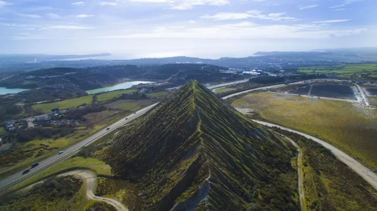 Aerial photograph of the Sky Tip in St Austell, Cornwall — a steep, pyramid-shaped china clay spoil heap surrounded by green fields, lakes, and the Cornish coastline in the distance.